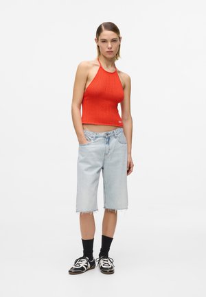 Kvinde iført en lys orange hæklet haltertop, lyse denim knælange shorts, sorte sokker og sort-hvide sneakers, stående med den ene hånd i lommen.