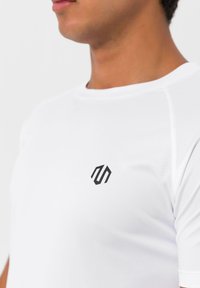 Hvid sports t-shirt med rund hals, korte ærmer og et lille sort logo på brystet. Glat, letvægtsstof.