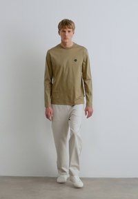 Világos olívazöld hosszú ujjú ing kerek nyakkivágással és kis logóval. Krémszínű, bő szabású nadrággal és fehér sportcipővel párosítva.