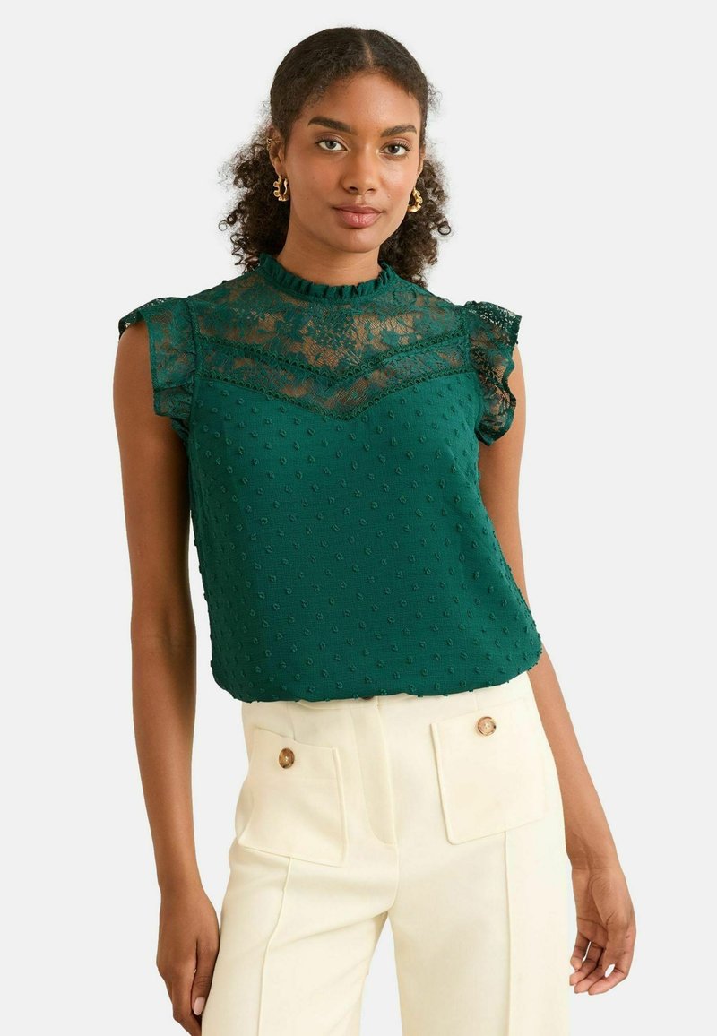 Blusa verde con sovrapposizione in pizzo sul collo, texture a pois e maniche a volant. Abbinata a pantaloni crema a vita alta.
