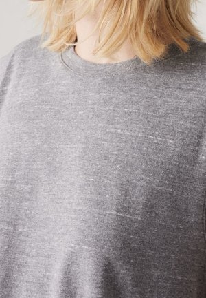 Blond persoon die een lichtgrijze, gespetterde trui met ronde hals draagt, het haar bedekt deels de nek, neutrale achtergrond.