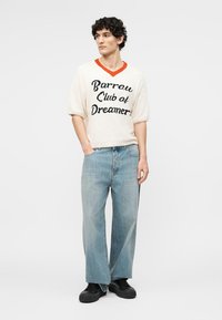 Νεαρός άνδρας με σγουρά μαλλιά, φοράει λευκό πουλόβερ με κοντά μανίκια με το κείμενο "Barrow Club of Dreamers", ανοιχτόχρωμο μπλε τζιν και μαύρα παπούτσια.