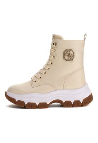 Stivaletti beige alla caviglia realizzati in materiale sintetico liscio, con suola in gomma bianca e marrone, design con lacci e un'emblema gioiello sul lato.