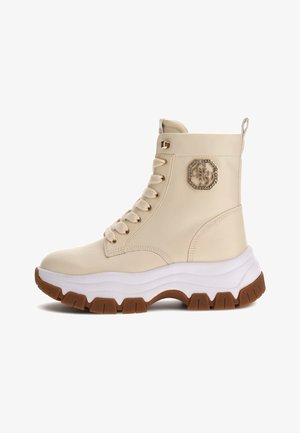 Stivaletti beige alla caviglia realizzati in materiale sintetico liscio, con suola in gomma bianca e marrone, design con lacci e un'emblema gioiello sul lato.