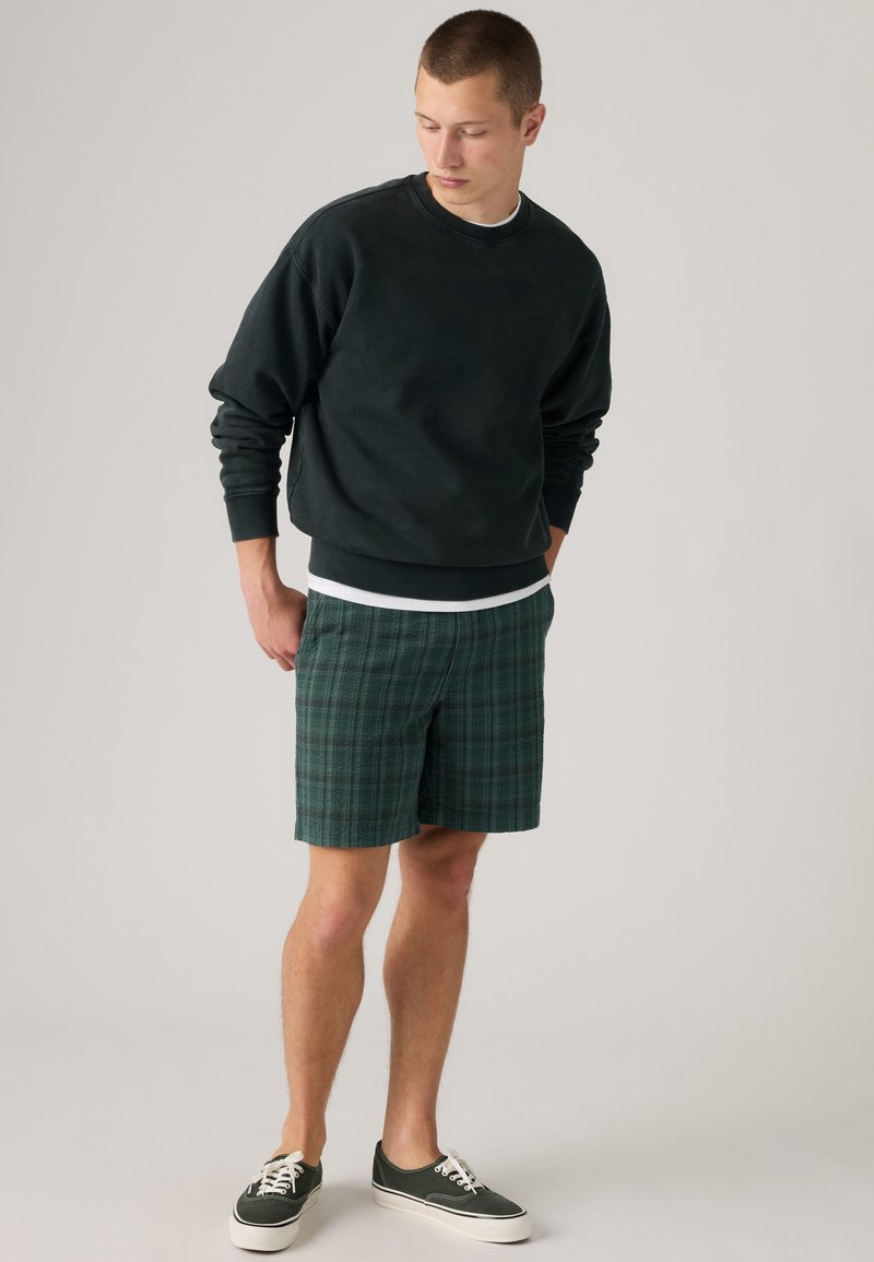 Giovane uomo che indossa una felpa nera, pantaloncini verdi a quadri e scarpe da ginnastica verde scuro, in piedi con le mani nelle tasche che guarda verso il basso.
