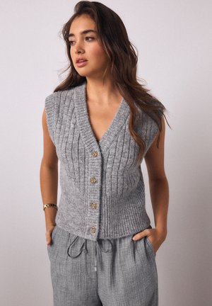 Fiatal nő hosszú barna hajjal, szürke ujjatlan, gombos kötött mellényt és laza szürke nadrágot visel, kezét a zsebében tartva.