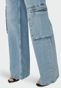 Svetlo modri denim cargo hlače s širokimi nogami, ki imajo stranske žepe, žepek z zadrgo in raztrgan rob. Nosijo se črne sandale z naramnicami.