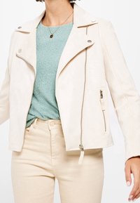 Jasnobeżowa sztuczna skórzana kurtka motocyklowa z srebrnymi elementami, zamykanymi kieszeniami oraz kołnierzem w kształcie litery V, połączona z fakturowanym turkusowym topem.