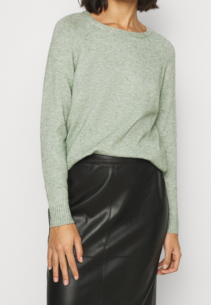 Femme portant un pull en maille vert clair rentré dans une jupe en cuir noire, debout devant un fond blanc uni.