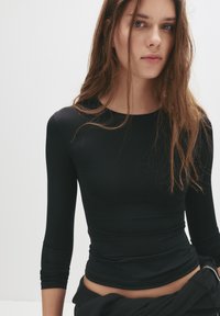 Schwarzes, figurbetontes Langarmoberteil aus glattem Material, mit rundem Halsauschnitt und leicht geraffter Textur an der Taille.