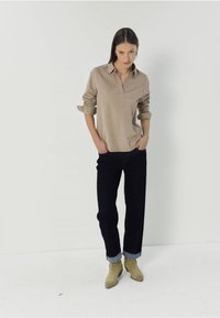 Polo a maniche lunghe beige con colletto, abbinato a jeans blu scuro arrotolati e stivaletti beige alla caviglia. Sfondo semplice.