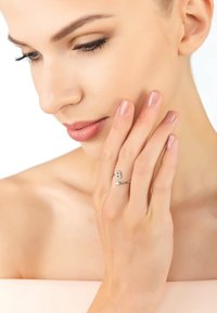 Anello d'argento con una decorazione a forma di "B" e due pietre trasparenti, indossato su una mano con unghie naturali su uno sfondo neutro.