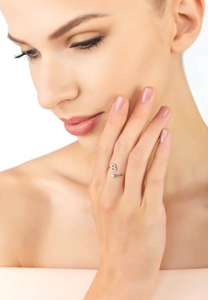 Silver ring featuring a decorative "B" and two clear stones, worn on a hand with natural nails against a neutral background.