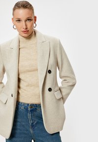 Blazer beige doppiopetto con bottoni neri, dotato di una coppia di tasche laterali, indossato sopra un dolcevita lavorato a maglia beige chiaro, completato da jeans blu.