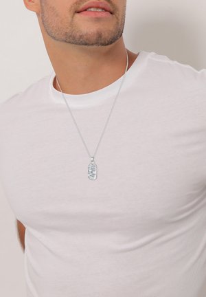 Collana con pendente in argento su una catena sottile, caratterizzata da un design rettangolare testurizzato, indossata su una maglietta bianca semplice.