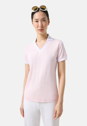 Vrouw draagt een lichtroze polo met korte mouwen en witte broek, met witte zonnebril op haar hoofd tegen een neutrale achtergrond.