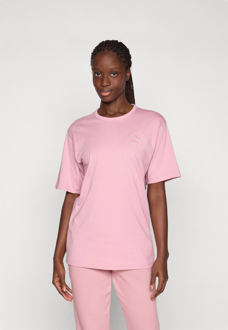 Rosa Baumwoll-T-Shirt mit rundem Halsausschnitt und kurzen Ärmeln, das ein kleines goldfarbenes Logo auf der oberen linken Brust besitzt.