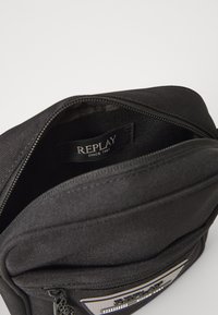 Black zippered bag with a fabric texture, featuring a prominent "REPLAY" tag inside. Additional zippered pocket visible on the front.
