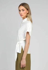 Jeune femme aux longs cheveux blonds portant un blouse blanche à manches courtes nouée à la taille et un pantalon vert olive, se tenant de profil devant un fond uni.