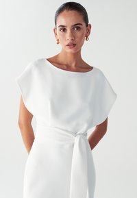 Bijela haljina s okruglim izrezom i širokim rukavima, s pojasom koji se veže. Glatka tkanina s lepršavom siluetom i minimalističkim dizajnom.