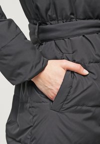 Musta eristetty takki, jossa on teksturoitu viimeistely, kiristetty vyötärö ja sivutasku, johon käsi lepää.