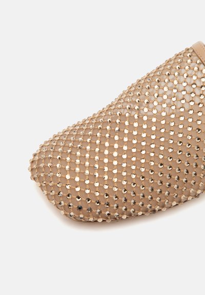 Gros plan sur un bout de chaussure beige recouvert d'un tissu en mesh orné de petites clous métalliques espacés de manière régulière.