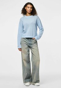 Helles blaugraues Strickpullover mit strukturiertem Muster und gewelltem Saum, kombiniert mit hellen, weit geschnittenen Jeans und weißen Schlupfschuhen.