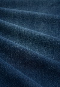 Zbliżenie na tkaninę z ciemnoniebieskiego denimu, pokazujące fakturę i diagonalne zagięcia w delikatnym oświetleniu.