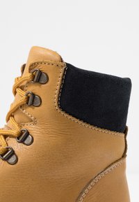 Stivale alla caviglia in pelle marrone con collo in pelle scamosciata nera, occhielli metallici rotondi e lacci gialli. Materiale strutturato e cuciture a contrasto visibili.