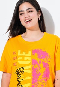 Camiseta oversize de color amarillo brillante con un gráfico de texto en negrita en rosa y negro; presenta una ilustración de un león y un cuello redondo. Tela de algodón suave.