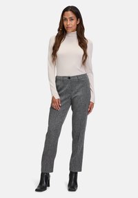 Helles Beige, langärmliger Rollkragenpullover, kombiniert mit grauen strukturierten, maßgeschneiderte Hosen und schwarzen Stiefeletten, stehend vor einem weißen Hintergrund.