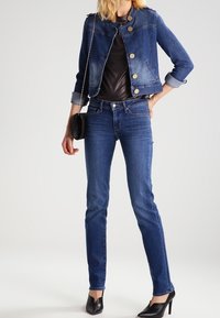 Denim-takki keskipunaisena, jossa on istuva muotoilu, napitus, ja käännetyt hihansuut, yhdistettynä tummansinisiin tiukkoihin farkkuihin ja mustiin korkokenkiin.