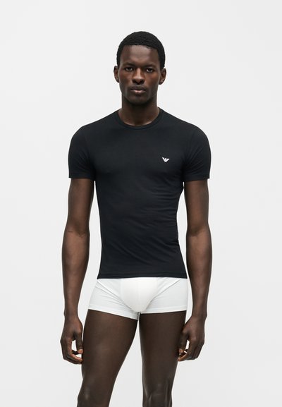 Άνδρας που φοράει εφαρμοστό μαύρο T-shirt και λευκά μποξεράκια, στέκεται μπροστά σε απλό ανοιχτό γκρι φόντο.