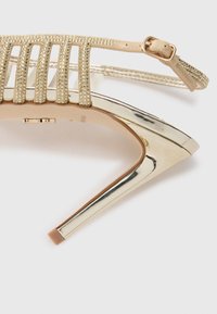 Sandali con tacco alto e cinturini, caratterizzati da una punta e un tacco in oro metallico, con cinturini texturizzati beige adornati con piccole pietre preziose.