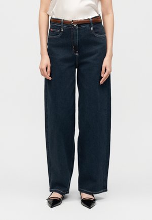 Donna che indossa jeans blu scuro a gamba larga con cintura marrone, top bianco e ballerine nere con punta a punta, in piedi davanti a uno sfondo neutro.