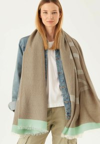 Écharpe tissée marron clair avec un motif géométrique, dotée d'une bordure à franges rayées vert menthe. Portée sur une veste en jean et une chemise blanche.