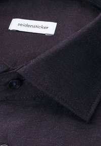 Camicia in tessuto scuro con colletto classico, chiusura con un singolo bottone nero e un'etichetta bianca cucita con la scritta "seidensticker" in testo nero.