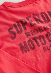 Röd bomullsskjorta med lång ärm som har en mörk, djärv grafisk tryck med texten "SUPERDRY RIDERS MOTO G" över ryggen i en slitna stil.