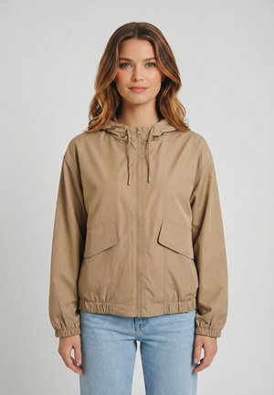 Giovane donna che indossa una giacca beige con cappuccio e cerniera, tasche a pattina frontali e jeans azzurri, in piedi contro uno sfondo chiaro e semplice.
