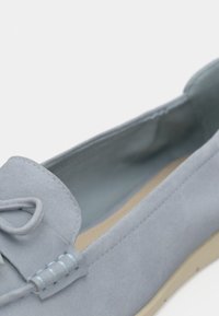 Mocassin en suède bleu clair avec une finition texturée, détail en nœud à l'avant et semelle beige. L'intérieur présente une doublure lisse de couleur bleu clair.