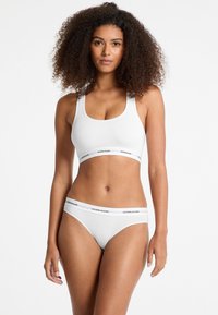Witte sportbeha en bijpassende bikini onderkant hebben een gladde textuur, met zwarte "GUESS JEANS" branding op de elastische banden.