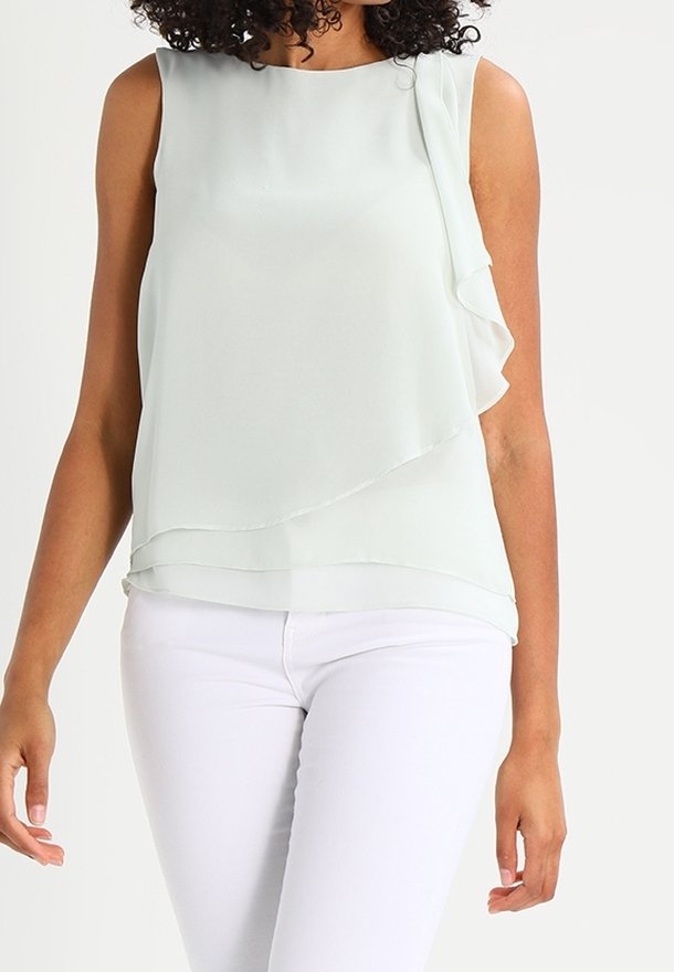 Femme portant un chemisier blanc sans manches à couches associé à un pantalon blanc ajusté sur un fond uni.