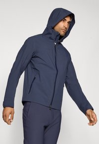 Navy waterproof jacket with a fitted design, adjustable hood, and zippered pockets. Smooth texture and minimal hardware accents.