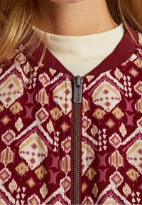 Bordeaux kleurige zip-up jack met geometrische patronen in crème, roze en geel. Heeft een staande kraag en een metalen ritsdetail.