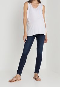 Mujer vestida con una blusa blanca sin mangas, jeans ajustados oscuros y sandalias planas beige, de pie contra un fondo claro y liso.