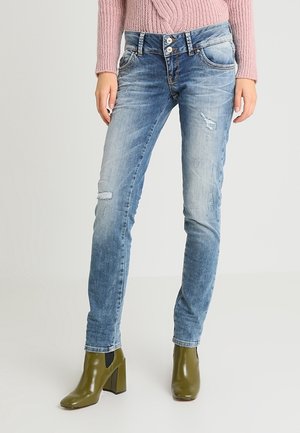 Femme portant un jean skinny bleu clair délavé, un pull en maille rose et des bottines à talons vert olive, debout sur un fond uni.
