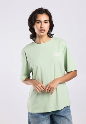 Woman with short dark hair wearing a light green oversized T-shirt and blue jeans, standing against a plain light background.