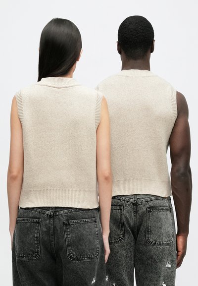 Μπεζ γιλέκο πλεκτό πουλόβερ χωρίς μανίκια με ριμπ λαιμόκοψη και τελείωμα, συνδυασμένο με φαρδιά σκούρα τζιν παντελόνια με διακοσμητικό ραφές.
