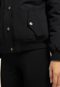 Giacca imbottita nera con bottoni a pressione, orlo a coste e tasca laterale con un logo discreto. Il materiale sembra liscio e isolante.