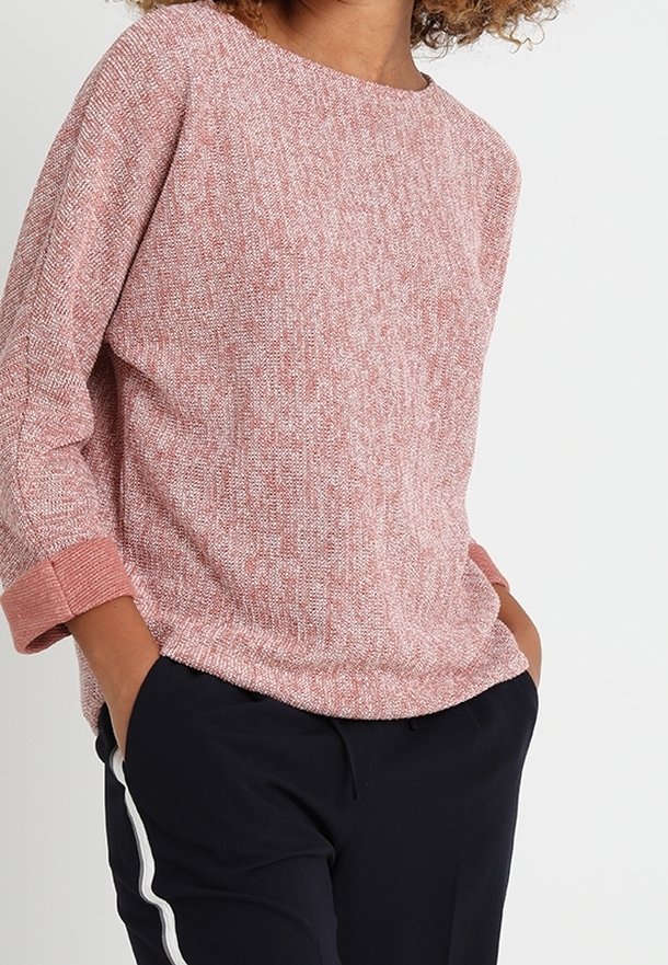 Femme portant un pull en maille texturé rose avec les poignets retroussés et un pantalon noir avec des bandes blanches sur les côtés, mains dans les poches, sur fond blanc.